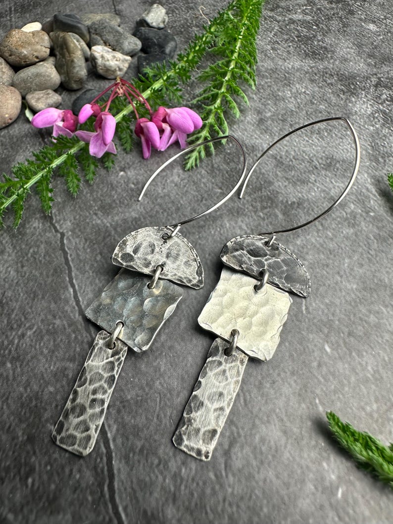 May include: A pair of silver dangle earrings with a hammered texture. Each earring features a half-circle top, a square middle, and a rectangular bottom, all connected by small loops. The earrings hang from curved silver wires. The background includes pink flowers and greenery.