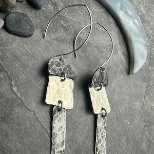 May include: Silver-toned, handcrafted earrings with a layered design. Each earring features three textured, geometric shapes suspended from a curved wire. The earrings have a hammered metal finish, creating a unique, artistic look. The earrings are displayed on a dark gray surface.