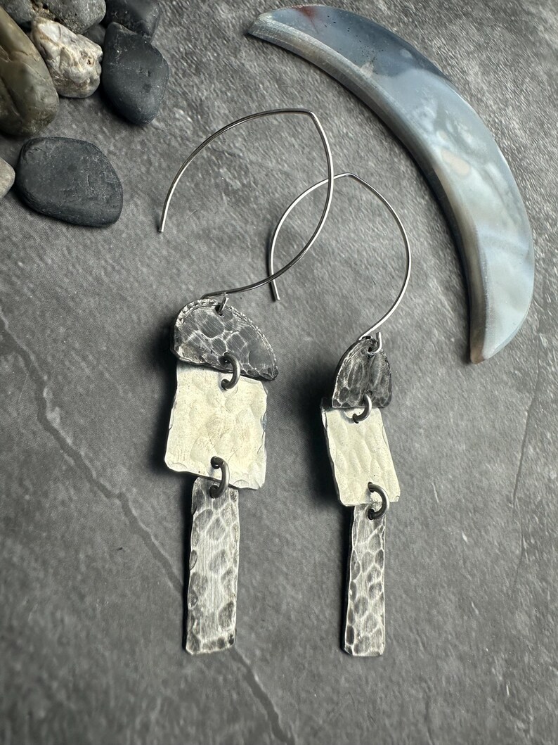 May include: Silver-toned, handcrafted earrings with a layered design. Each earring features three textured, geometric shapes suspended from a curved wire. The earrings have a hammered metal finish, creating a unique, artistic look. The earrings are displayed on a dark gray surface.
