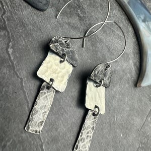 May include: A pair of silver earrings with a modern, geometric design. Each earring features three textured, rectangular elements in varying shades of silver and gray, connected by small metal rings. The earrings have a hook closure.