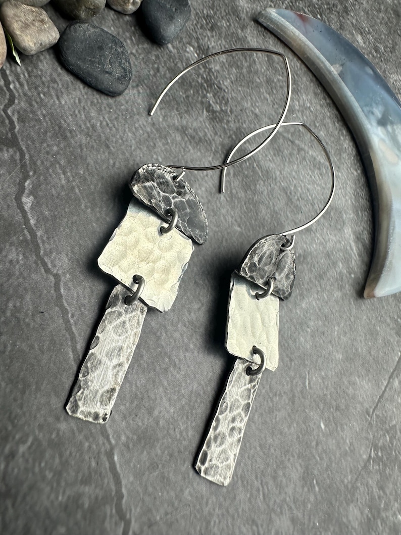 May include: A pair of silver earrings with a modern, geometric design. Each earring features three textured, rectangular elements in varying shades of silver and gray, connected by small metal rings. The earrings have a hook closure.
