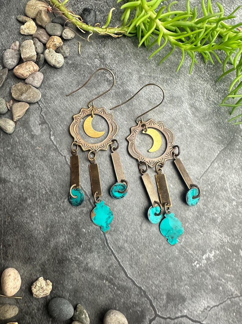 May include: A pair of dangle earrings with a celestial theme. Each earring features a brass sunburst design with a gold crescent moon, rectangular brass bars, and turquoise-colored teardrop-shaped accents. The earrings have a hook closure.