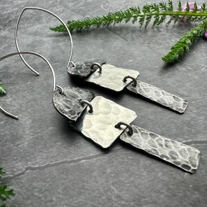 May include: Silver-toned dangle earrings with a hammered texture. Each earring features a curved wire hook and three connected, textured metal shapes: a heart, a square, and a rectangle. The earrings are displayed on a dark gray surface with greenery.