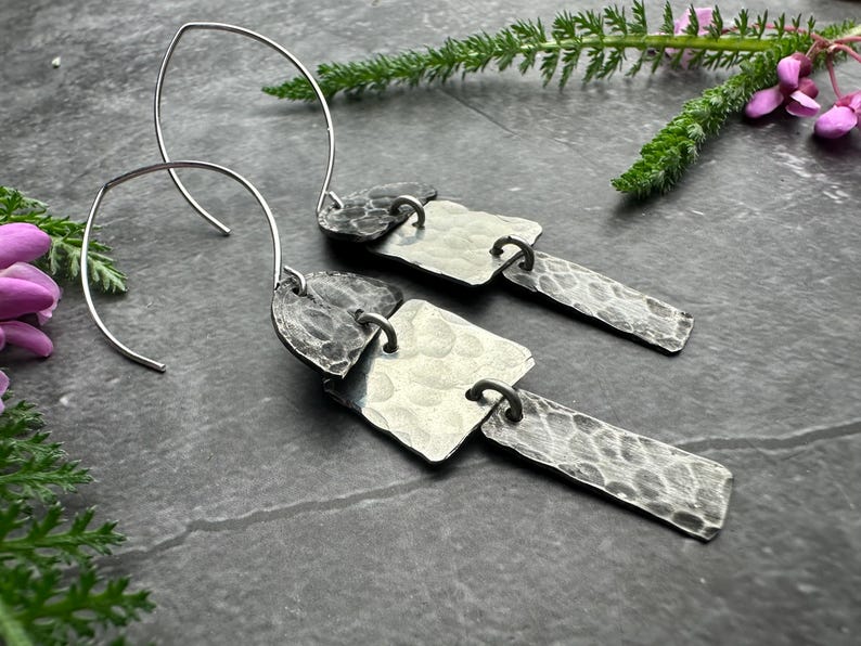 May include: Silver-toned dangle earrings with a hammered texture. Each earring features a curved wire hook and three connected, textured metal shapes: a heart, a square, and a rectangle. The earrings are displayed on a dark gray surface with greenery.