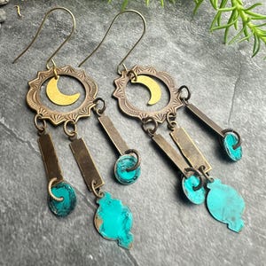 May include: A pair of handcrafted earrings with a celestial theme. Each earring features a brass sunburst design with a gold crescent moon, rectangular brass bars, and turquoise-colored accents. The earrings have a hook closure.