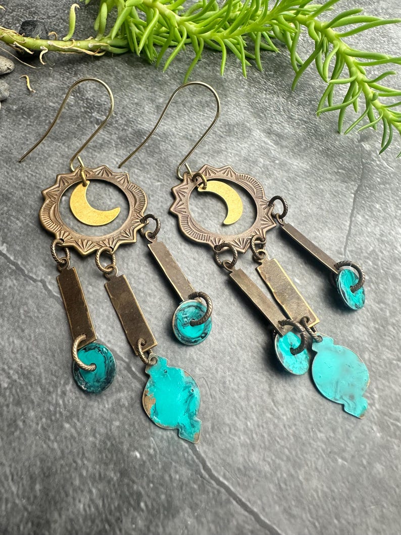 May include: A pair of handcrafted earrings with a celestial theme. Each earring features a brass sunburst design with a gold crescent moon, rectangular brass bars, and turquoise-colored accents. The earrings have a hook closure.