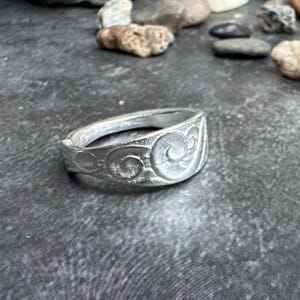 May include: A silver ring with an embossed swirl design. The ring is made of a light-colored metal and has a flat band. The ring is displayed on a textured gray surface with small rocks in the background. Jewelry.