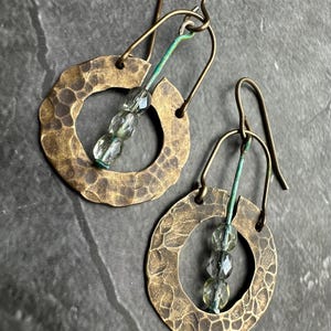 May include: Handmade earrings with textured brass hoops and green wire accents. Each earring features three clear, faceted beads. The ear wires and hoops have a rustic, antique brass finish.