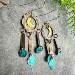 May include: A pair of handcrafted earrings with a celestial theme. Each earring features a brass sunburst frame with a gold crescent moon, rectangular metal bars, and turquoise teardrop-shaped accents. The earrings hang from thin, gold-colored ear wires.