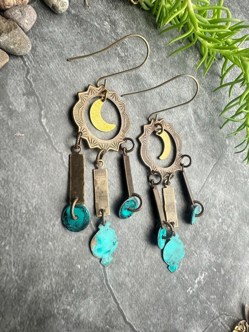 May include: A pair of handcrafted earrings with a celestial theme. Each earring features a brass sunburst frame with a gold crescent moon, rectangular metal bars, and turquoise teardrop-shaped accents. The earrings hang from thin, gold-colored ear wires.