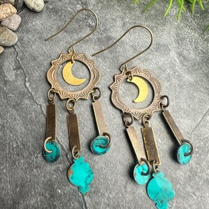May include: A pair of dangle earrings with a celestial theme. Each earring features a brass-colored sunburst design with a gold crescent moon in the center. Below hang rectangular and teardrop-shaped turquoise accents, connected by small metal links.