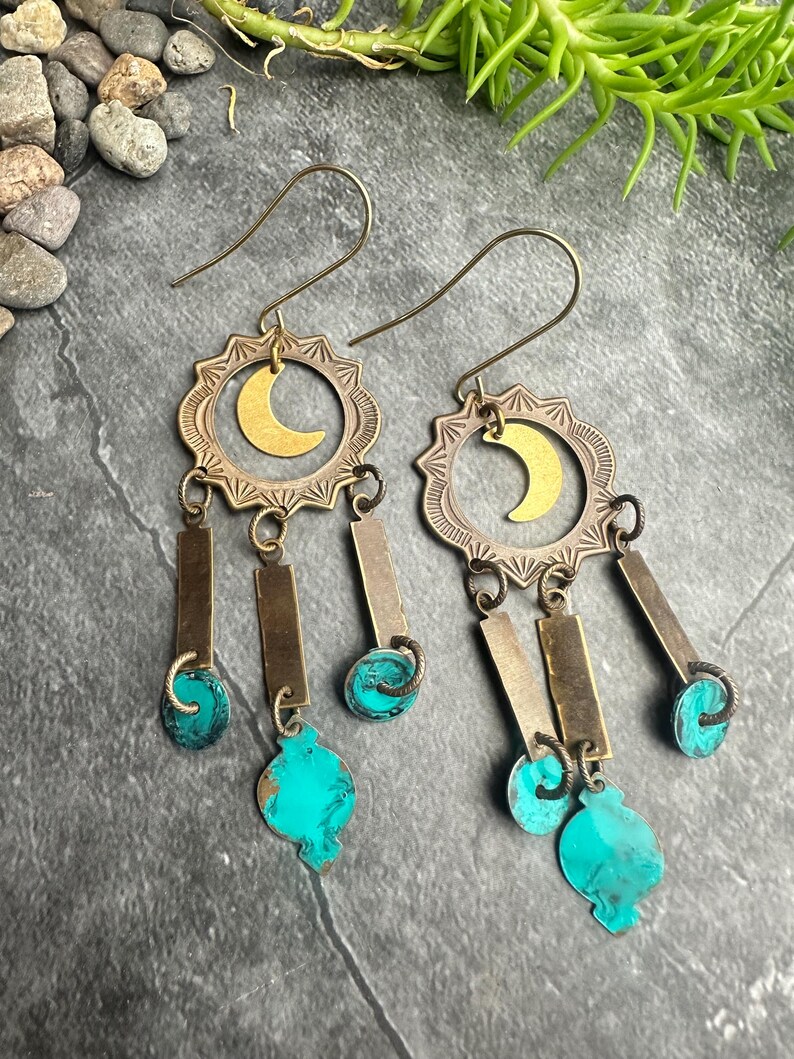 May include: A pair of dangle earrings with a celestial theme. Each earring features a brass-colored sunburst design with a gold crescent moon in the center. Below hang rectangular and teardrop-shaped turquoise accents, connected by small metal links.