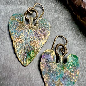 Pode incluir: Um par de brincos dourados com um design de folha. As folhas são texturizadas e têm um acabamento multicolorido, incluindo tons de verde, azul e roxo.