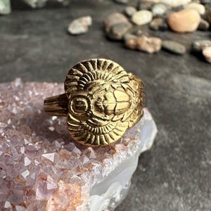 May include: A gold-colored ring with an intricate design featuring a central, stylized face with radiating details. The ring is set on a light purple crystal formation, with a dark background of pebbles and stones.