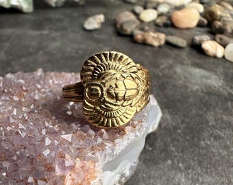 May include: A gold-colored ring with an intricate design featuring a central, stylized face with radiating details. The ring is set on a light purple crystal formation, with a dark background of pebbles and stones.