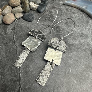 May include: A pair of silver earrings with a hammered texture. Each earring features a half-circle top, a square middle, and a rectangular bottom, all connected by small loops. The earrings have a dark, oxidized finish and dangle from thin, curved ear wires.