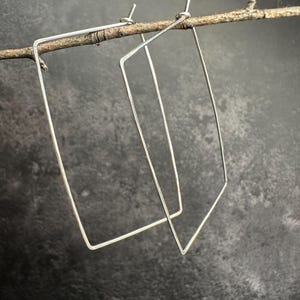May include: A pair of silver-toned, geometric rectangle hoop earrings. The earrings are suspended from a thin, natural-colored twig against a dark, textured background. The earrings have a minimalist, modern design.