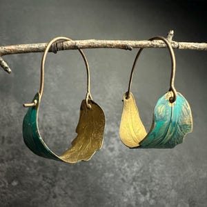 May include: A pair of handcrafted earrings with a feather design. The earrings are made of gold-colored metal with a teal and gold finish. The earrings are suspended from a thin, curved wire.