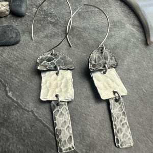 May include: A pair of silver earrings with a hammered texture. Each earring features a half-circle top, a rectangular middle piece, and a smaller rectangular bottom piece, all connected by small hoops. The earrings have a hook closure.