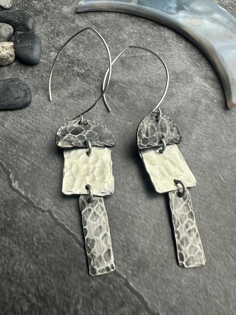 May include: A pair of silver earrings with a hammered texture. Each earring features a half-circle top, a rectangular middle piece, and a smaller rectangular bottom piece, all connected by small hoops. The earrings have a hook closure.