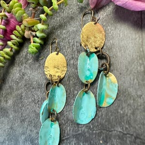 May include: Handmade earrings with a layered design. The earrings have a gold-coloured, hammered disc at the top, with turquoise-coloured, oval-shaped dangles. The earrings are connected with small metal rings and wire. The earrings are displayed with greenery and purple flowers.