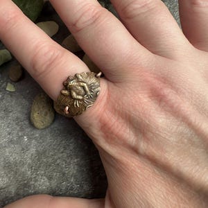 May include: A bronze-colored ring featuring a detailed frog design. The ring's focal point is a sculpted frog resting on a lily pad, showcasing intricate textures and a vintage aesthetic. The ring is worn on a finger.