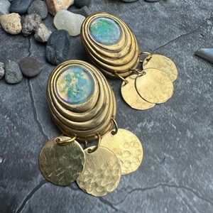 May include: A pair of gold-toned earrings. Each earring features an oval-shaped top with a central iridescent stone and a series of concentric ridges. Below, three hammered-finish, circular charms dangle from each earring. The earrings are displayed on a dark gray surface.