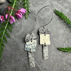May include: Artisan-crafted silver earrings with a hammered texture. Each earring features three geometric shapes: a half-circle, a square, and a rectangle. The earrings are suspended from a curved silver wire. The earrings are displayed on a dark gray surface.