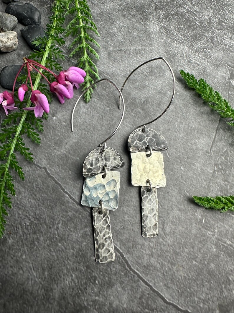 May include: Artisan-crafted silver earrings with a hammered texture. Each earring features three geometric shapes: a half-circle, a square, and a rectangle. The earrings are suspended from a curved silver wire. The earrings are displayed on a dark gray surface.