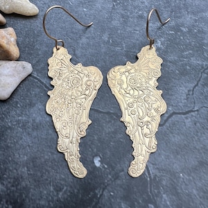 Może przedstawiać: Para złotych kolczyków w kształcie skrzydeł. Każdy kolczyk ma skomplikowany, wytłoczony wzór z kwiatowymi i wirującymi wzorami. Kolczyki mają zapięcie na haczyk i są prezentowane na ciemnym, teksturowanym tle.