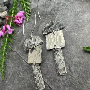 May include: A pair of silver earrings with a hammered texture. Each earring has a half-circle top, a square middle piece, and a rectangular bottom piece. The earrings hang from a curved wire.