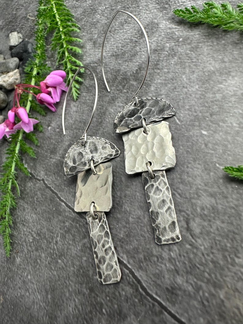 May include: A pair of silver earrings with a hammered texture. Each earring has a half-circle top, a square middle piece, and a rectangular bottom piece. The earrings hang from a curved wire.