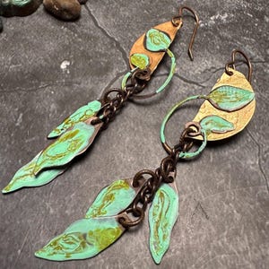 May include: A pair of long, dangling earrings with a green and gold leaf design. The earrings are made of metal and have a chain link connecting the top and bottom parts.