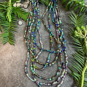 May include: A selection of beaded necklaces in shades of blue, green, purple, and gold. The necklaces are composed of small, round beads and are layered. The backdrop is a dark grey surface with green foliage, creating a natural aesthetic.