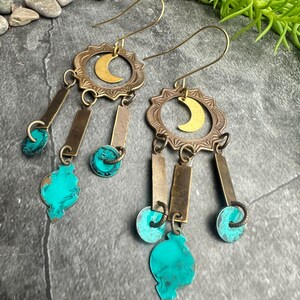 May include: A pair of brass earrings with a crescent moon design. The earrings feature turquoise-colored accents and rectangular and circular brass elements. The earrings have a bohemian style.