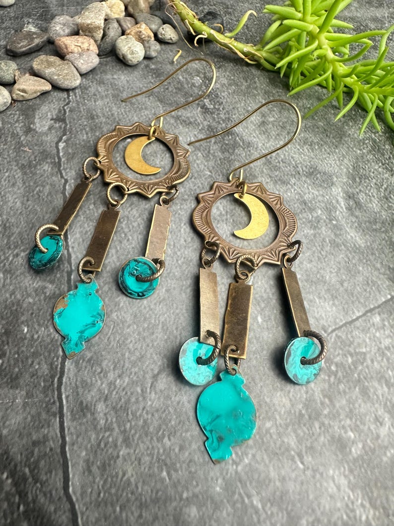 May include: A pair of brass earrings with a crescent moon design. The earrings feature turquoise-colored accents and rectangular and circular brass elements. The earrings have a bohemian style.