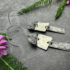 May include: A pair of silver earrings with a hammered texture. Each earring features a curved wire hook, a square, and a rectangular element connected by small rings. The earrings are displayed on a dark surface with greenery and pink flowers.