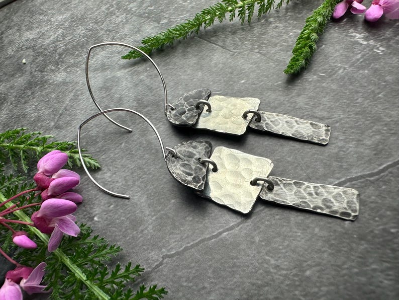 May include: A pair of silver earrings with a hammered texture. Each earring features a curved wire hook, a square, and a rectangular element connected by small rings. The earrings are displayed on a dark surface with greenery and pink flowers.