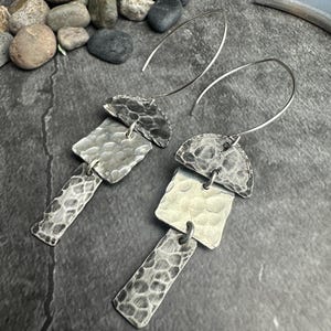 May include: A pair of silver earrings with a hammered texture. Each earring features a half-circle top, a square middle, and a rectangular bottom, all connected by small loops. The earrings hang from curved ear wires.