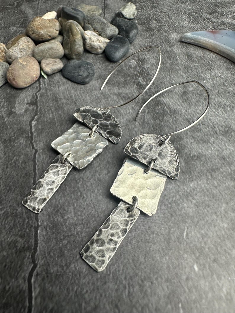 May include: A pair of silver earrings with a hammered texture. Each earring features a half-circle top, a square middle, and a rectangular bottom, all connected by small loops. The earrings hang from curved ear wires.