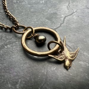 May include: A bronze-toned necklace featuring a circular pendant with a small, dark pearl and a detailed bird charm. The chain is a matching bronze color, and the pendant has a hammered texture. The necklace is displayed on a dark, textured surface.