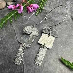 May include: A pair of silver earrings with a hammered texture. Each earring has a half-circle top, a square middle, and a rectangular bottom, all connected by small loops. The earrings hang from a curved wire. The background includes flowers and greenery.