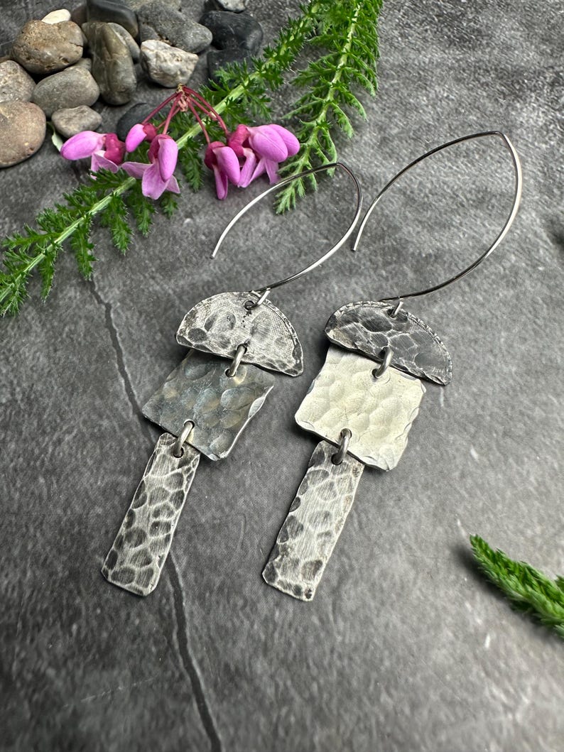 May include: A pair of silver earrings with a hammered texture. Each earring has a half-circle top, a square middle, and a rectangular bottom, all connected by small loops. The earrings hang from a curved wire. The background includes flowers and greenery.
