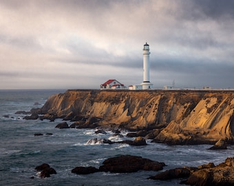 May include: A white lighthouse stands tall on a rocky cliff overlooking a vast ocean. The sky is cloudy and the water is choppy.