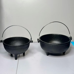 May include: Two black cast iron cauldrons with rounded bowls, three short legs, and thin silver-colored metal handles. The cauldrons are positioned side-by-side, featuring a textured exterior finish and open tops, showcasing a functional design.