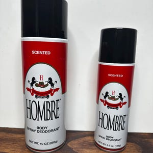 Op de afbeelding: Twee spuitbussen Hombre body spray deodorant. De bussen zijn rood en wit met zwarte tekst en een logo met een hoed en paarden. De grotere bus is 283g, en de kleinere is 156g.