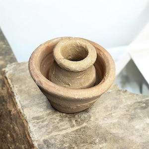 May include: A small, unglazed terracotta pot with a rounded base and a smaller, cylindrical pot nested inside. The outer pot has a wide, shallow bowl shape. The color is a warm, earthy brown. The pots appear to be handmade.
