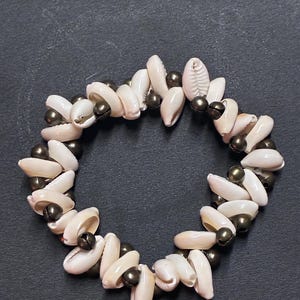 May include: A circular bracelet constructed from alternating off-white cowrie shells and small, dark metallic spherical beads. The shells are arranged in a repeating pattern, creating a textured, layered appearance around the entire circumference.