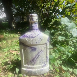 May include: A decorative, off-white bottle with purple marbling. The bottle has a rectangular shape with a short neck and a textured surface. The word "BISHOP" is visible on the front of the bottle. The bottle is outdoors, surrounded by green foliage.