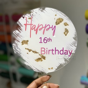 以下が含まれることがあります： ゴールドの斑点と「Happy 16th Birthday」の文字が入った白いアクリル製のケーキトッパーです。